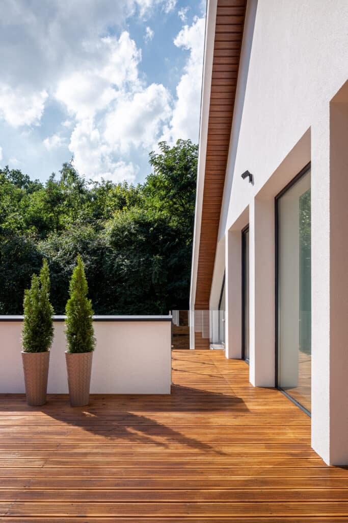 Terrasse moderne en bois clair avec deux plantes vertes en pots sur un fond de façade blanche, grandes fenêtres et forêt luxuriante sous ciel bleu nuageux.