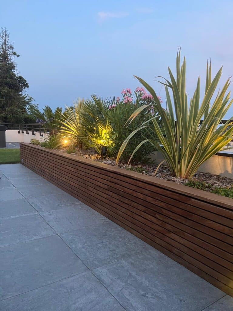 Jardinière en bois avec plantes exotiques éclairées par des spots jaunes, au crépuscule, sur une terrasse carrelée grise.